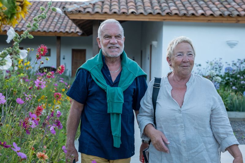 Smiling older couple standing among colorful flowers. Senior man and woman in casual clothes outdoors. Happy retirees enjoying nature and leisure activities.