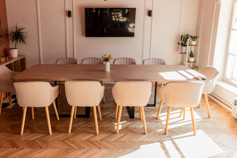 Sunlight streaming through the window illuminates a modern meeting room, highlighting the large wooden table, comfortable chairs, and sleek TV screen, creating a welcoming and productive atmosphere