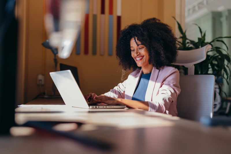 Young cheerful african american businesswoman sitting at desk typing on laptop, working in modern office enjoying natural sunlight, smiling