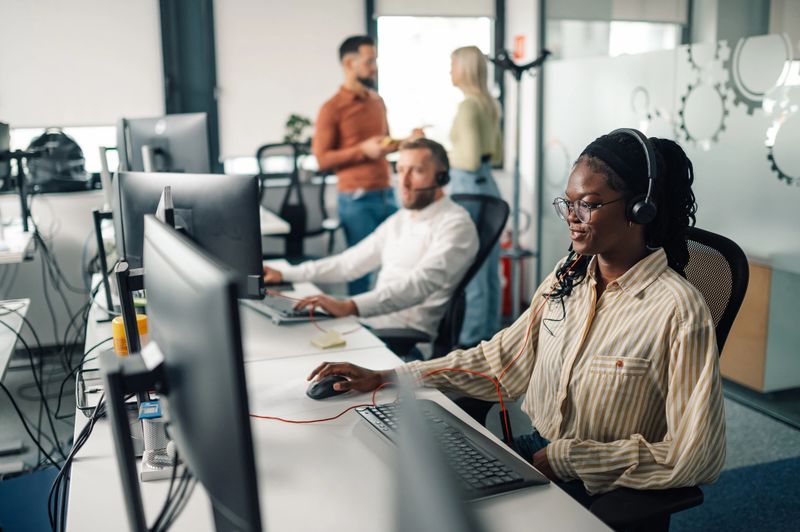 Black female support agent wearing headset is using computer while working in a call center with her colleagues in the background