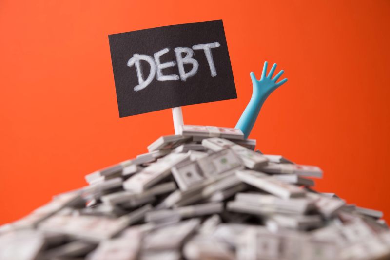 Pop out hand from heap US dollar banknotes and DEBT label white wall background. Over burdened from loan high interest rate, bankruptcy, past due payment, stress in economy crisis and so on concept.