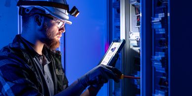 Electrician wearing safety gear working on an electrical panel with a tablet.