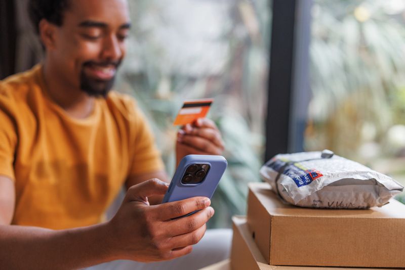 A man wearing a casual outfit holds a credit card while using a smart phone. Boxes and packages are visible, emphasizing the concept of online shopping at home and secure payment in e-commerce.