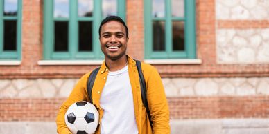 Young man smiling with a soccer ball and backpack outside a building.