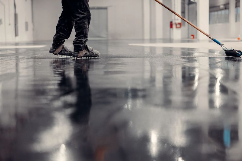 Worker applying epoxy coating on the industrial floor with a roller, reflecting surface, in a modern warehouse setting.