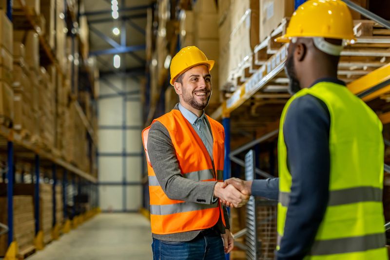 Two warehouse workers greet each other with a handshake while wearing safety vests and helmets. The image represents teamwork, professionalism, and productivity in a storage or logistics environment.