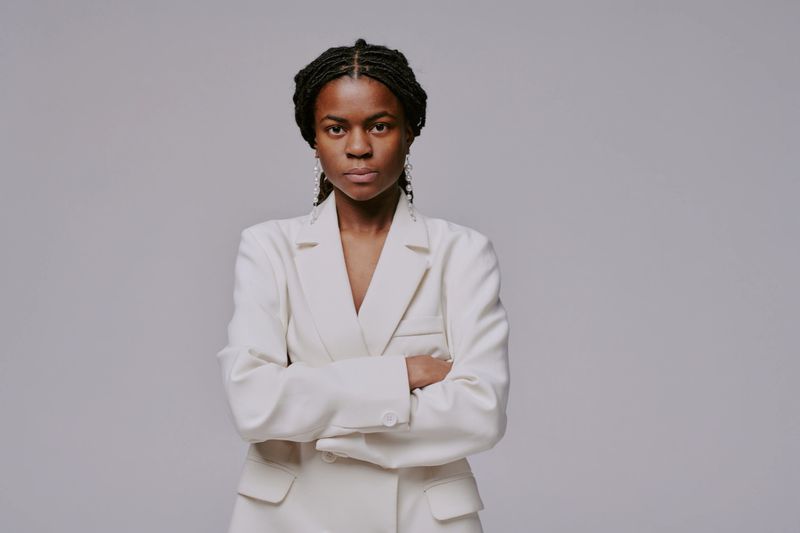 Portrait of confident Black woman crossing arms while standing against gray background in formal white blazer, showing determination and professionalism