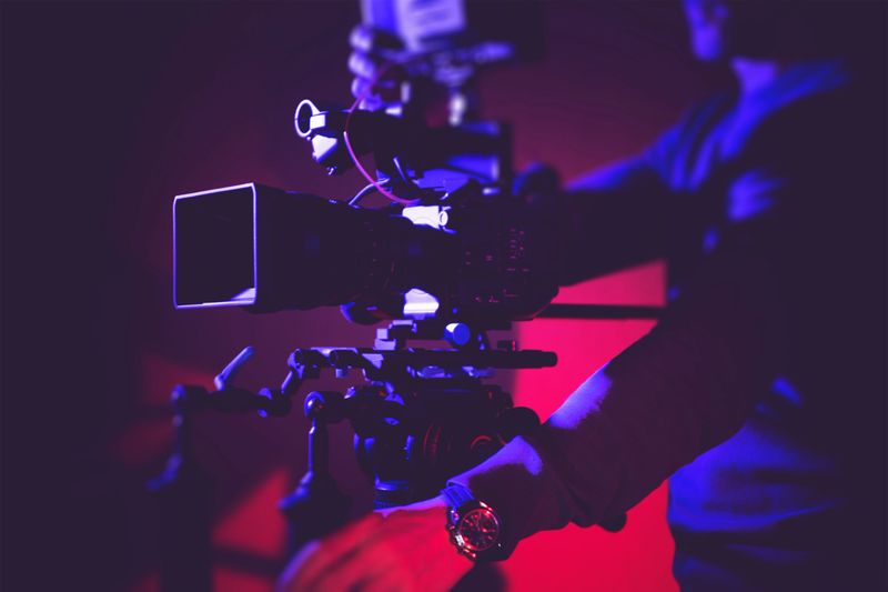 A camera operator carefully adjusts filming equipment with colorful lighting in the background, focused on achieving the perfect shot.