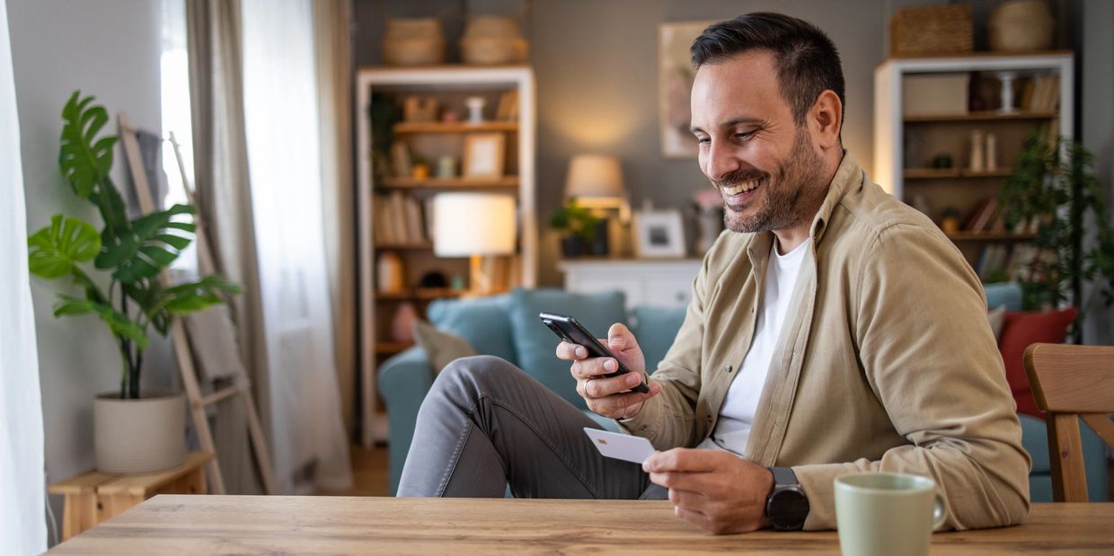 Smiling man using phone and holding credit card at home.