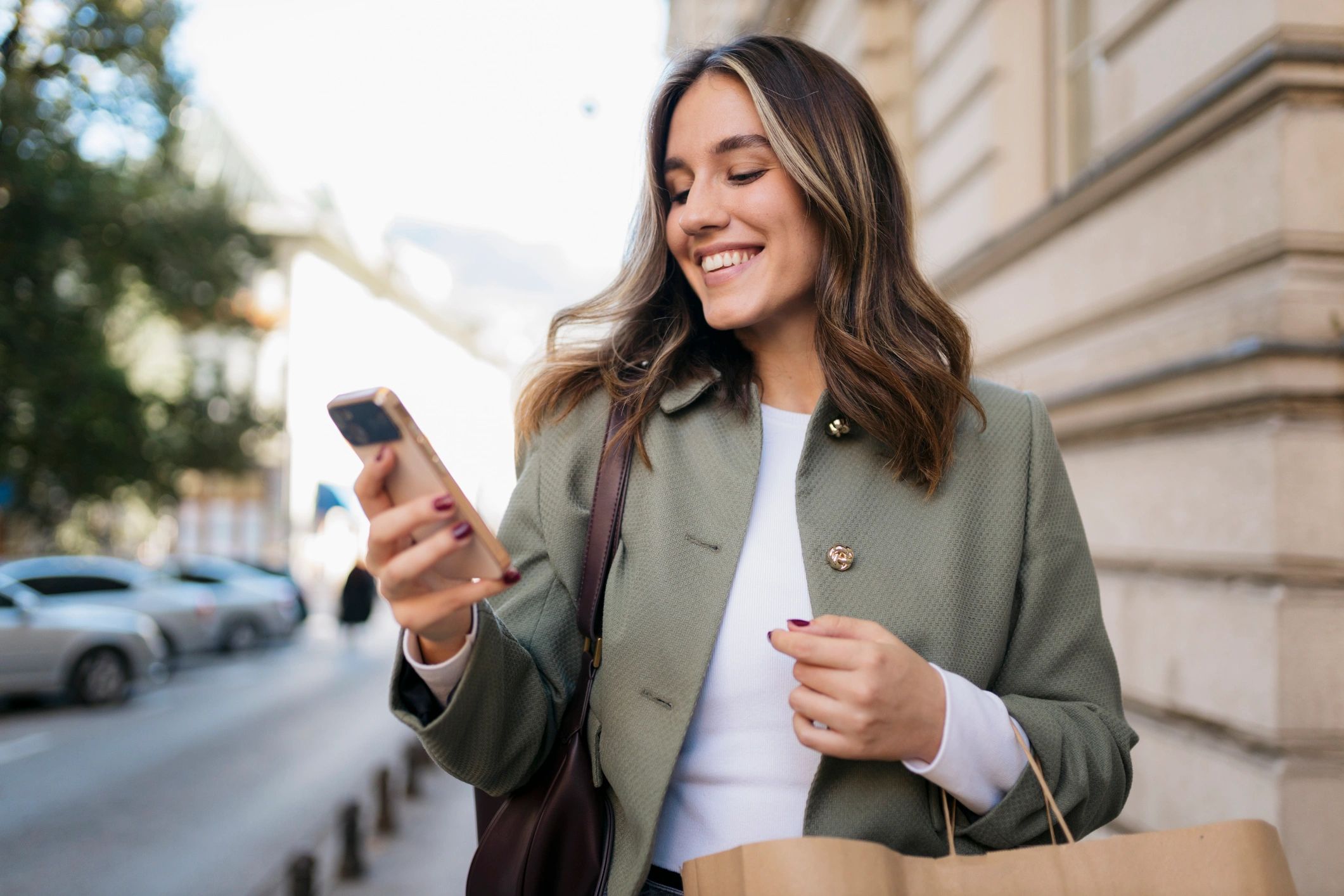Fashionable young woman smiles while using her smartphone and carrying a shopping bag on a city street