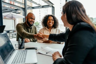 Two people shaking hands with a woman across a desk in an office.