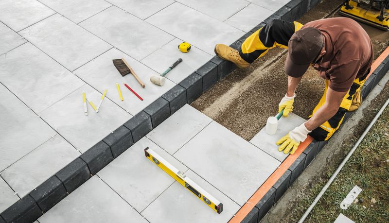 A skilled contractor is carefully positioning pavers in a backyard patio area, using tools like a level and measuring tape to ensure precision in the installation process.