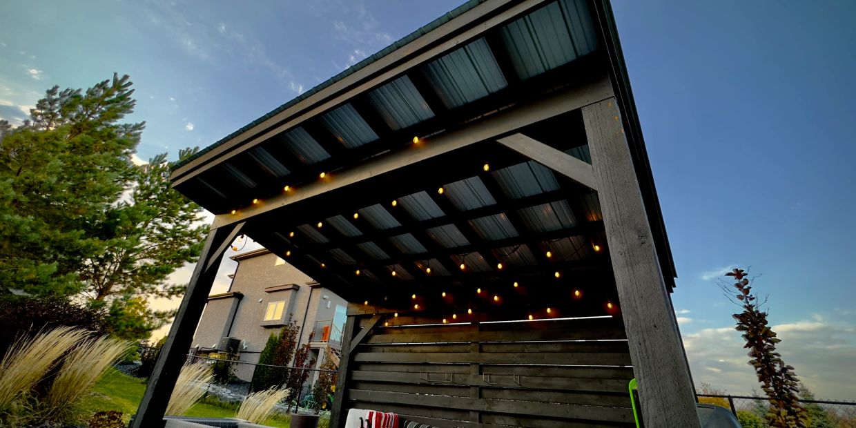 A wooden pergola with string lights in a backyard at dusk.