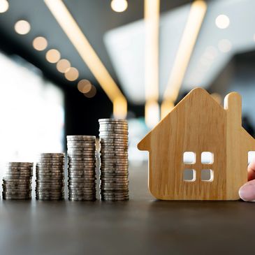 Stacked coins increasing in size next to a wooden house held by a hand.