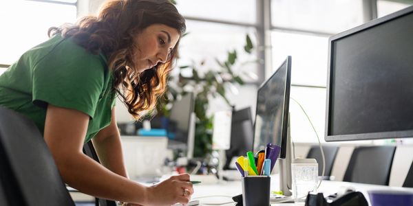 Woman working and writing notes at desk with computer monitors.