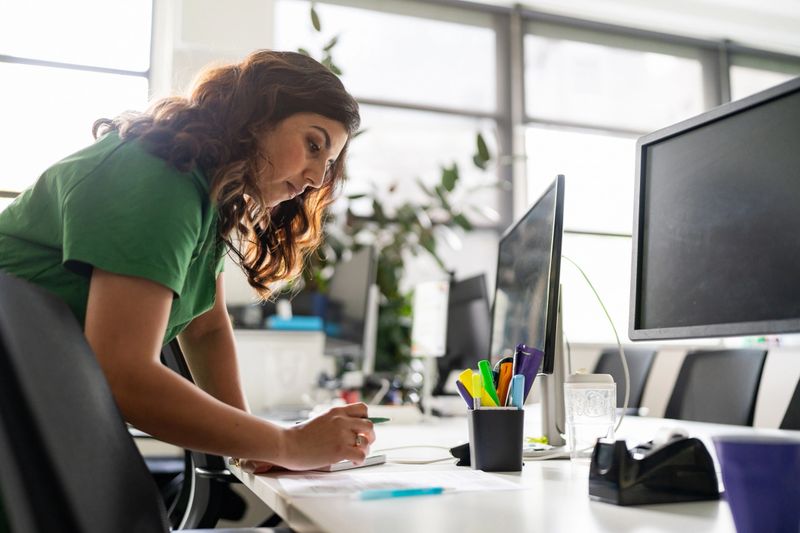 A focused professional woman working on documents at an office desk, showcasing dedication and productivity in a modern workspace environment, surrounded by office tools and natural light.