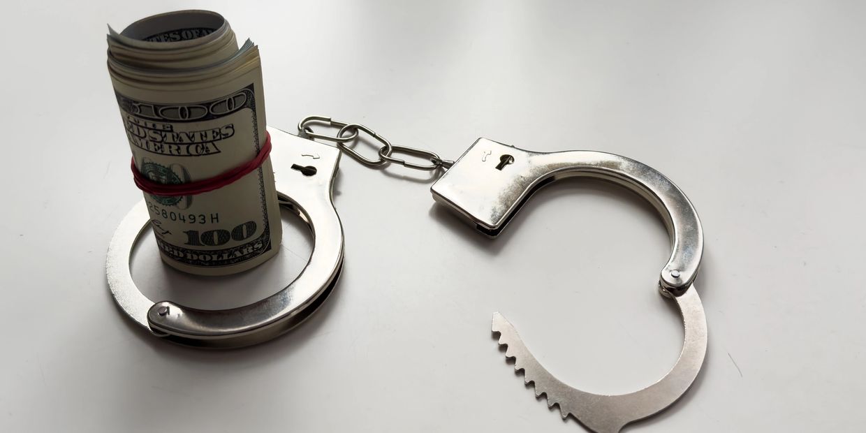 Rolled hundred-dollar bills secured in one handcuff, with the other handcuff open on a white surface.