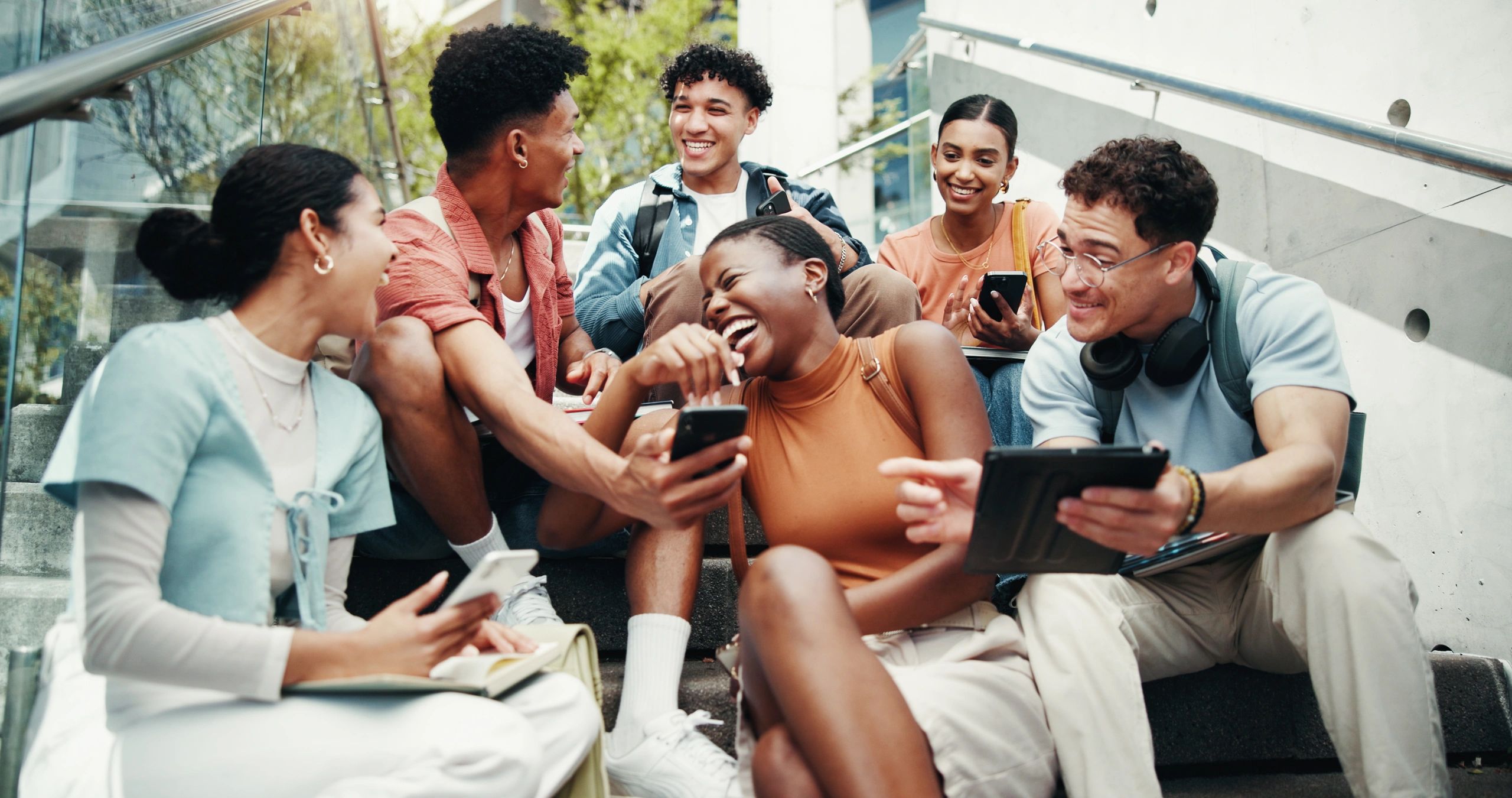 Group of young people sitting on some outside steps, laughing and looking at phones and ipads.