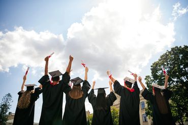 Graduates in caps and gowns celebrate outdoors, holding diplomas high against a bright sky.