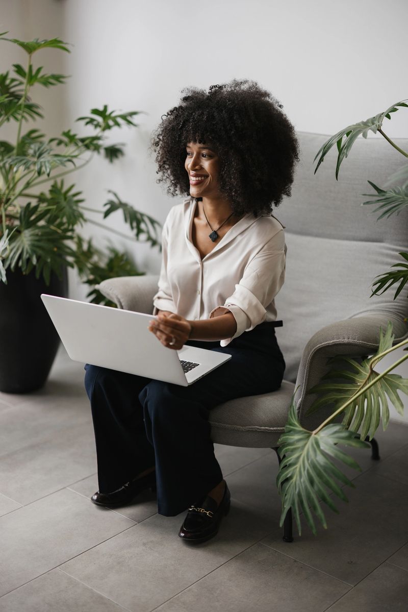 An engaged woman joyfully using her laptop in a stylish chair is surrounded by leafy green plants, creating an inviting atmosphere of innovation and inspiration.