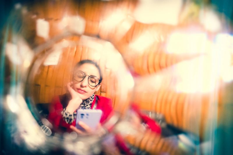 A young businesswoman uses her smartphone, sitting relaxed in a restaurant. The image is captured through a drinking glass, creating a bizarre effect. This image highlights the unique Lomo photography/Lomography style.