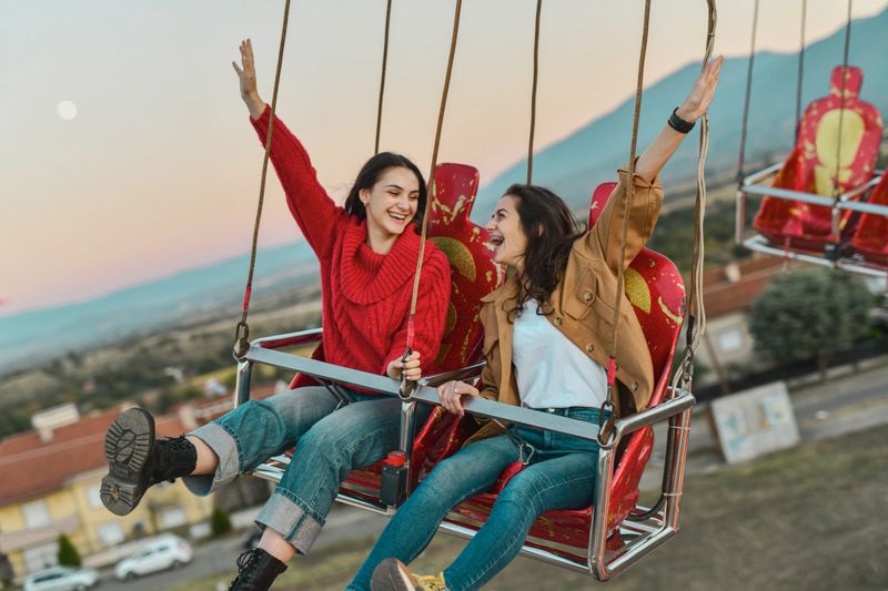 Beautiful Female Friends Laughing Loudly Enjoying Sunset Chain Swing Ride at Amusement Park