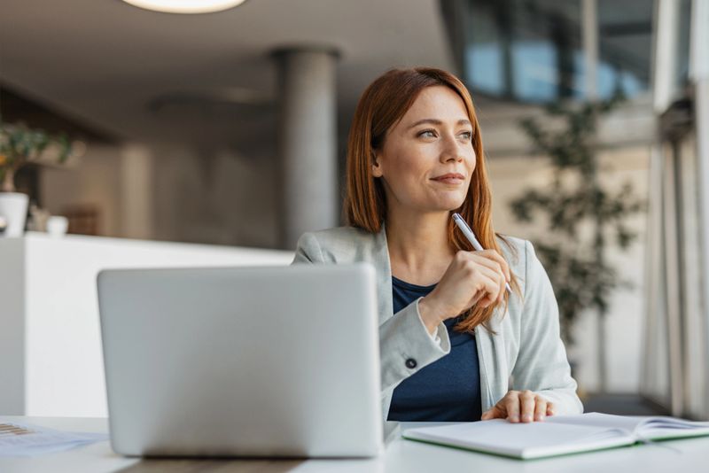 A pensive businesswoman gazes thoughtfully out the window, contemplating ideas and strategies in her chic office, emphasizing a blend of professionalism and introspection.
