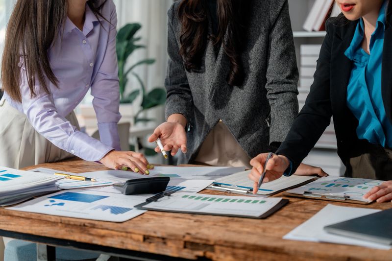 Three businesswomen are working together, analyzing financial charts and graphs, discussing company strategy, and collaborating on a project to achieve success