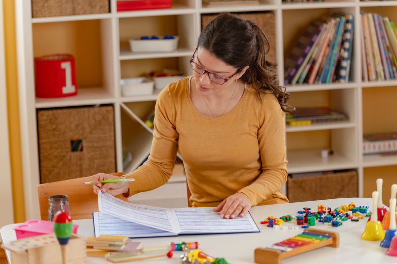 A preschool teacher in a mustard sweater reviews a lesson plan in a notebook, surrounded by educational toys and materials in a well-organized classroom