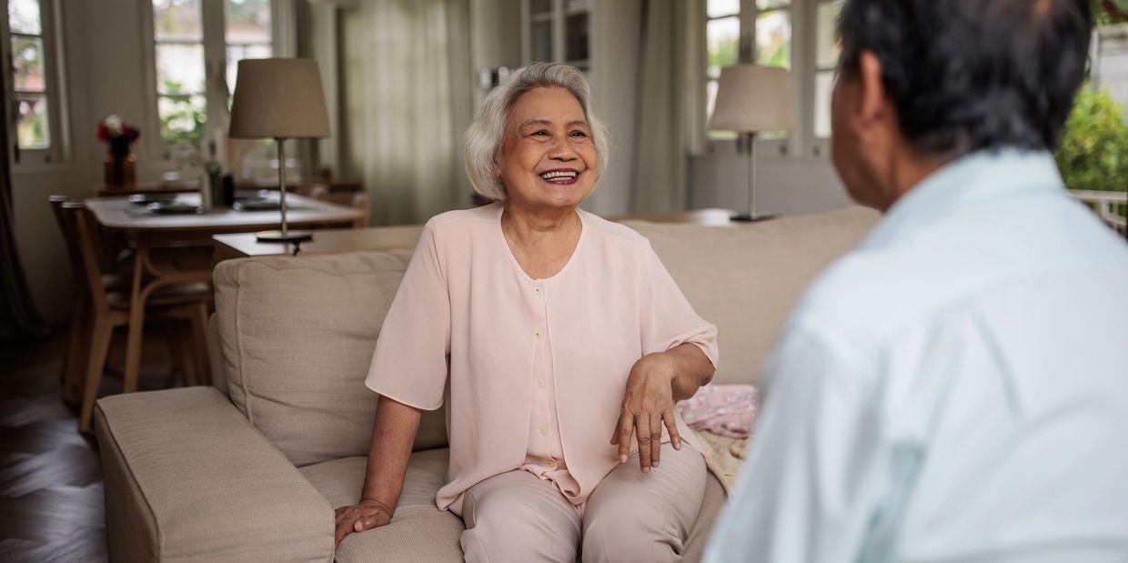 Elderly woman smiling while sitting on a couch, engaged in conversation with a man.