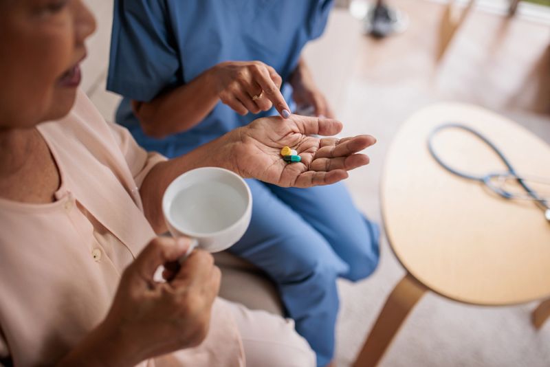 Compassionate female caregiver is assisting an elderly Thai woman in taking her medication while sitting comfortably on a sofa.