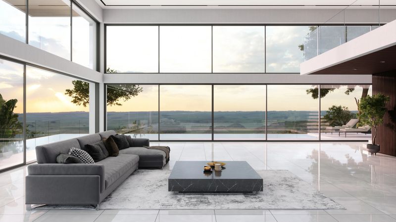 A modern living room featuring floor-to-ceiling windows, a gray sectional sofa, and a low-profile coffee table. The open-concept space is filled with natural light and offers a panoramic view of the surrounding landscape.
