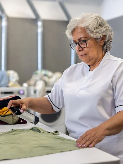 Senior woman ironing a green shirt in a laundry setting.