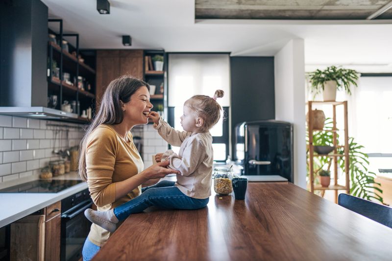 Mother and toddler sharing a joyful moment in a modern kitchen, with the child feeding the mother while both smile amidst sleek appliances and indoor plants