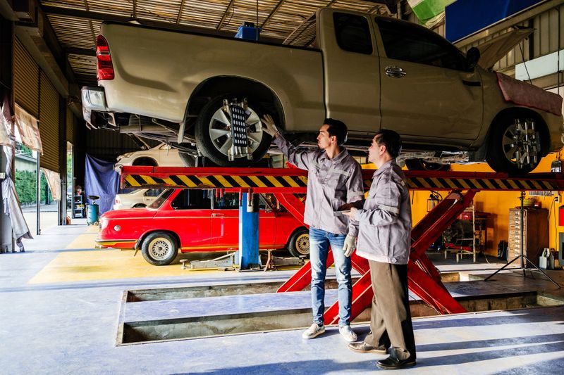 Asian mechanics inspecting a pickup truck on hydraulic lift at auto repair shop. Ideal for concepts like vehicle maintenance, wheel alignment, diagnostics, car service, garage work, mechanical repair