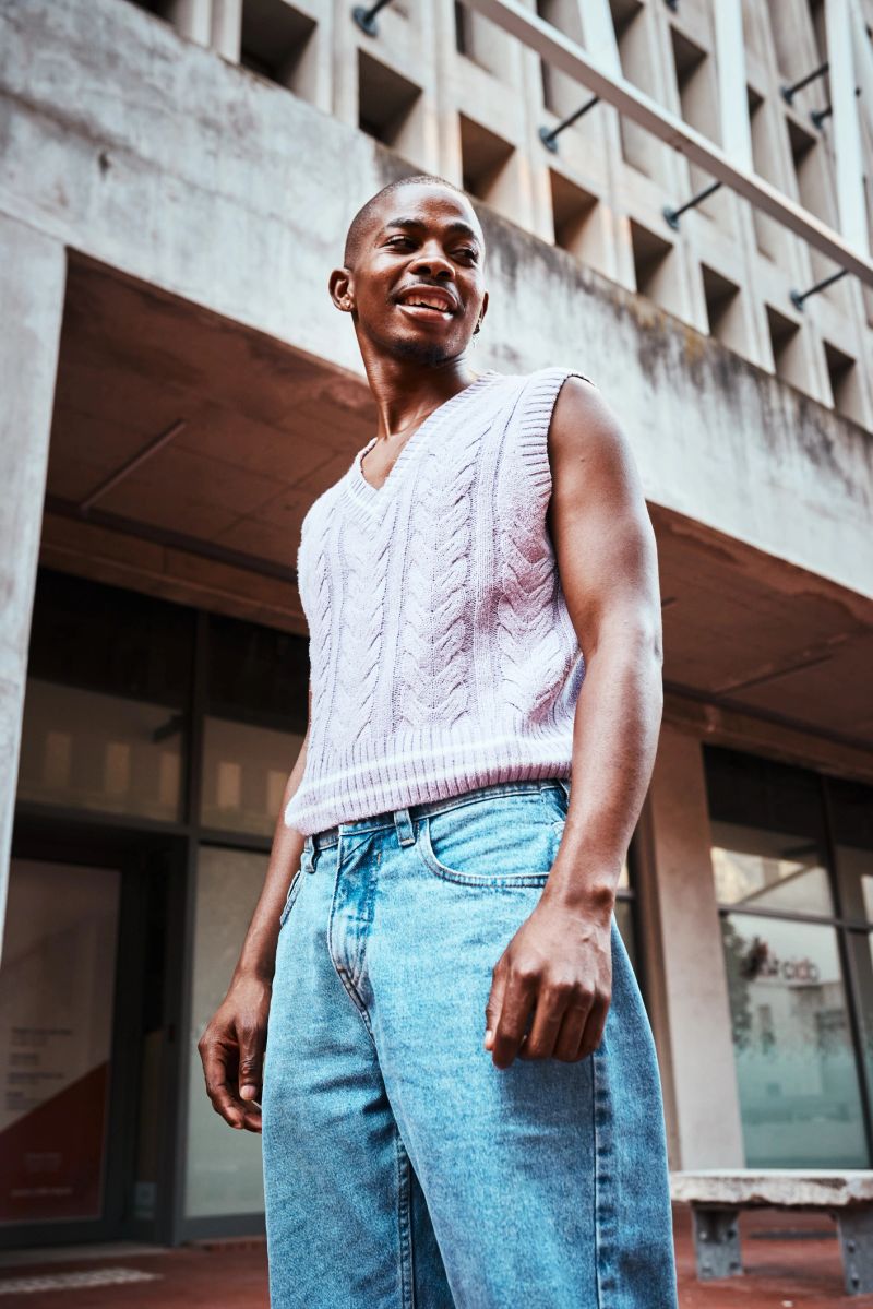 A confident young man wearing a knitted pullover tank top and jeans, smiling effortlessly. Photographed in a modern urban setting with a stylish vibe and natural light.