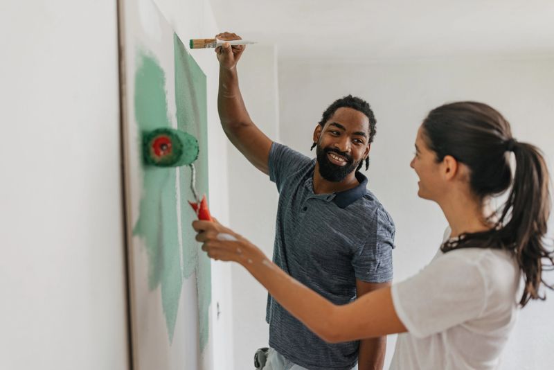A cheerful couple is collaborating while painting, capturing a moment filled with laughter and creativity, showcasing their commitment to a shared home project.