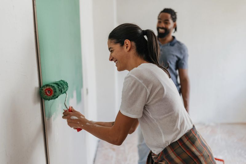 A smiling woman engages in painting a wall alongside her partner, capturing joy, creativity, and the shared experience of home renovation with a vibrant green palette.