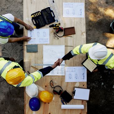 Two construction workers shaking hands over a table with blueprints and tools.