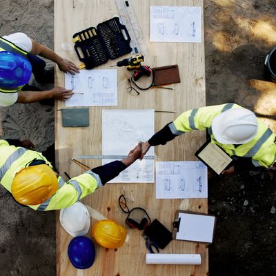 Three contractors in hard hats shaking hands over blueprints during a project planning meeting