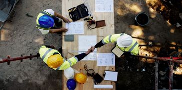 Two construction workers shaking hands over a table with blueprints and tools.