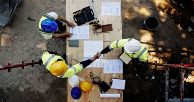 Two construction workers shaking hands over plans at a worksite.