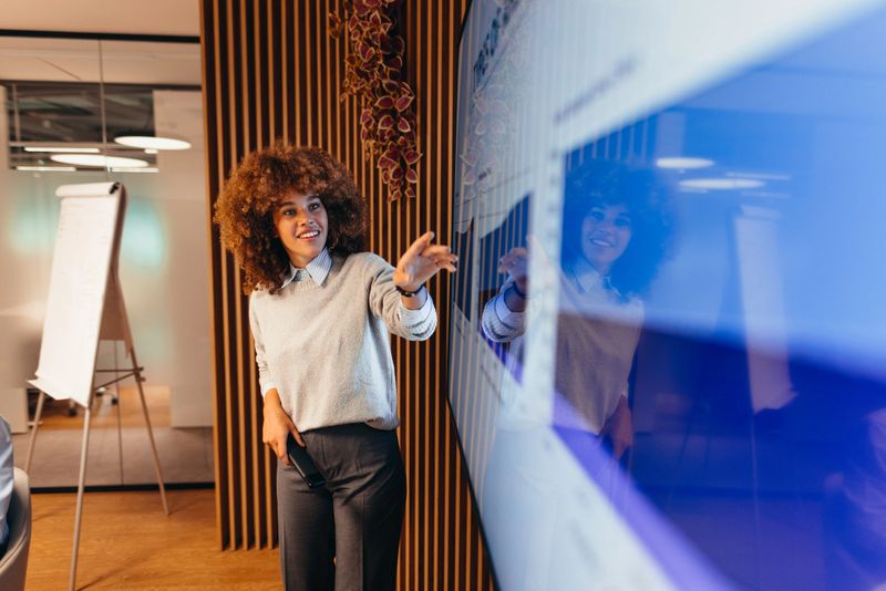 Young businesswoman engaging colleagues with a large interactive monitor while illustrating a presentation in a modern office setting, fostering collaboration and communication