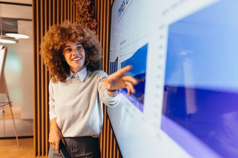 Young businesswoman with curly hair is pointing at a graph displayed on an interactive whiteboard, delivering a presentation in a modern office environment