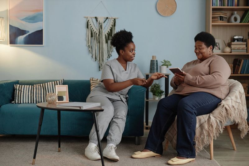 Full length of smiling African American elderly woman learning how to pay bills online using digital tablet with help of accommodating social worker, sitting together in living room