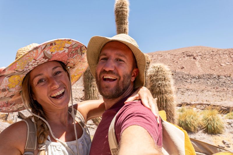 Smiling couple posing together in desert setting. Happy travelers exploring arid terrain. Tourists in wide-brimmed hats enjoying sunny outdoor adventure.