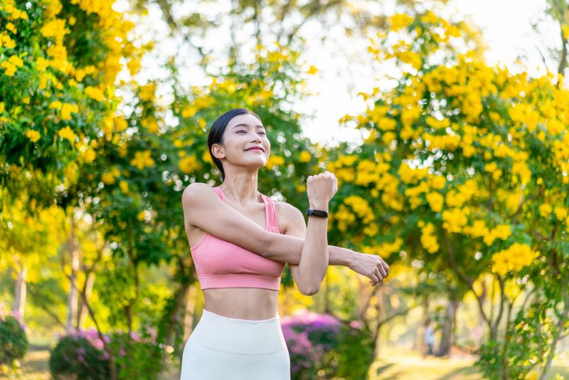 Young Asian Woman Stretching in a Green Park Surrounded by Beautiful Flowers on a Sunny Day