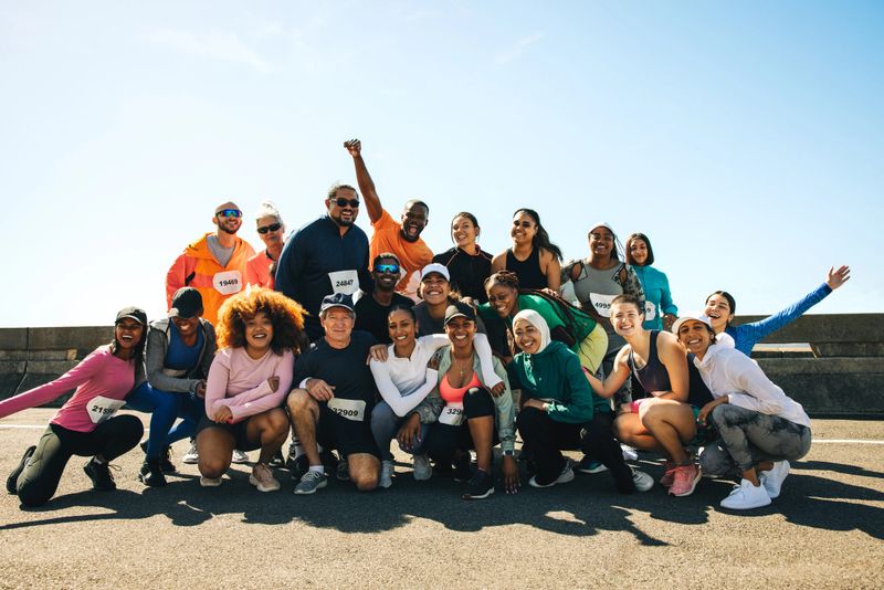 A diverse group of enthusiastic runners gather for a cheerful group photo under a clear blue sky. The running club, wearing colorful athletic gear and race numbers, exudes friendship and joy.
