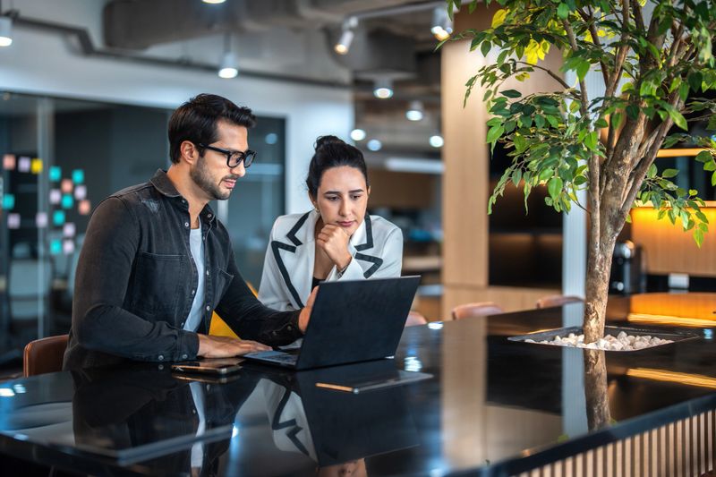 Two businesspeople talking and working on a laptop together at a desk in a modern office.