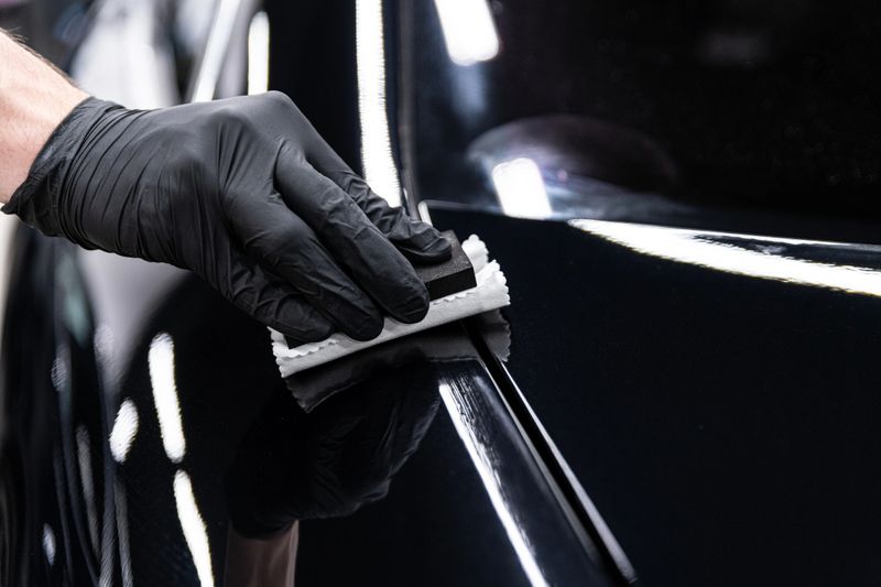 Car wash or detailing studio employee applies ceramic or graphene coating to black car paint ensuring deep gloss and advanced protection